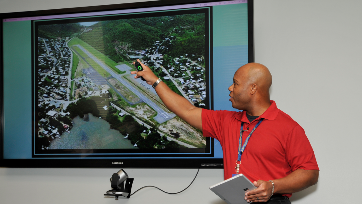 Man standing in front of a presentation of an island airport with one runway surrounded by mountains.